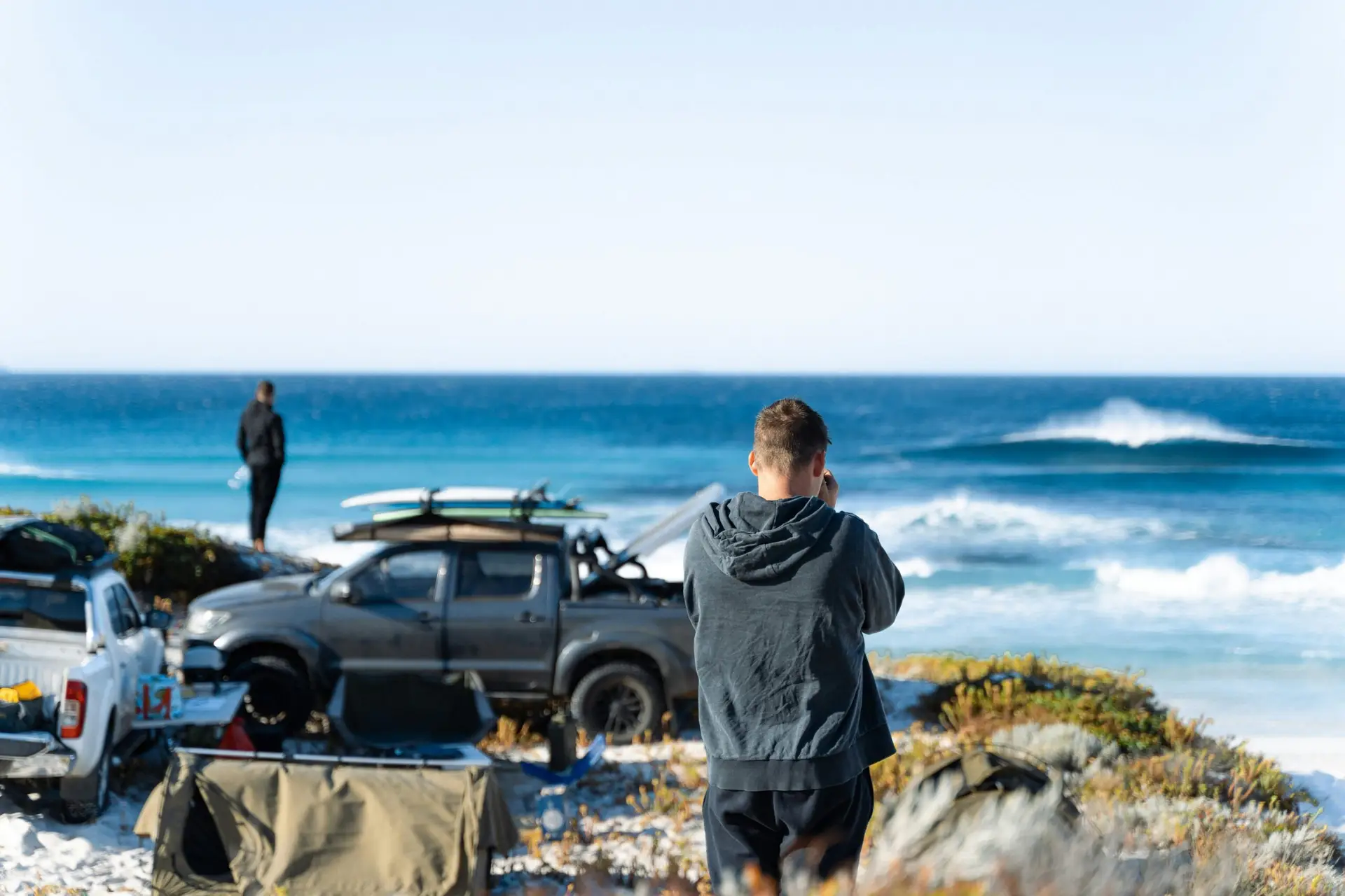 Two men enjoying a beach day with trucks, surfboards, and ocean waves.